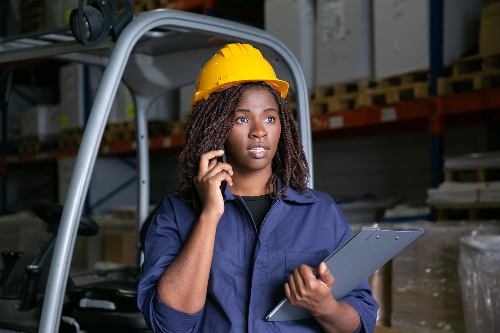 Focused Black warehouse worker in yellow hardhat standing near forklift and talking on cell. Shelves with goods in background. Medium shot. Labor or communication concept